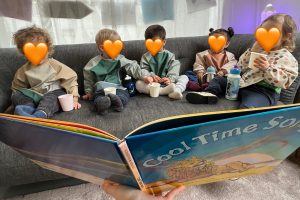 Fünf Kinder sitzen gemeinsam auf einem Sofa, schauen in ein aufgeschlagenes Buch und genießen kleine Snacks und Getränke in der Spielgruppe von Tiny Hands Creations