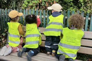 Vier Kinder mit Leuchtwesten sitzen draußen auf einer Bank, aufgenommen von hinten während des Feriencamps in Zürich