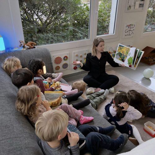 Acht Kinder sitzen in der Sofaecke und hören der Spielgruppenleiterin beim Vorlesen eines Buches in der Spielgruppe in Zürich zu.