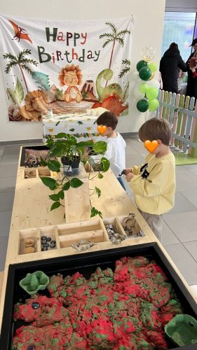 Zwei Kinder spielen am sensorischen Tisch mit kinetischem Sand und Pflanzen im Dino-Geburtstagsbereich von Tiny Hands Creations, Fotoleinwand im Hintergrund