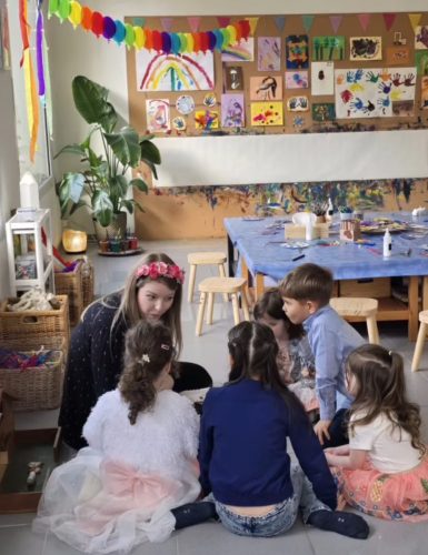 Fünf Kinder, einige als Feen verkleidet, sitzen und hören der Leiterin zu. Im Hintergrund sind Malwand und Basteltisch bei einer Märchenland-Geburtstagsparty von Tiny Hands Creations sichtbar.