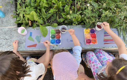 Drei Kinder malen mit Wasserfarben draußen neben Pflanzen bei Tiny Hands Creations in Zürich, frische Luft und natürliche Umgebung