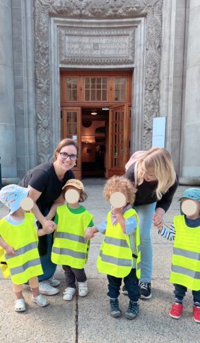 Zwei Lehrerinnen begleiten mehrere Kinder mit Leuchtwesten vor einem Museum in Zürich während eines Camps von Tiny Hands Creations