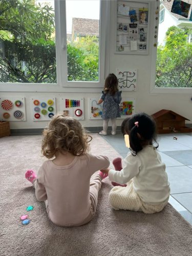 Zwei Kinder sitzen auf dem Teppich und spielen, ein weiteres Kind steht an den Aktivitätstafeln in der Spielgruppe in Zürich.