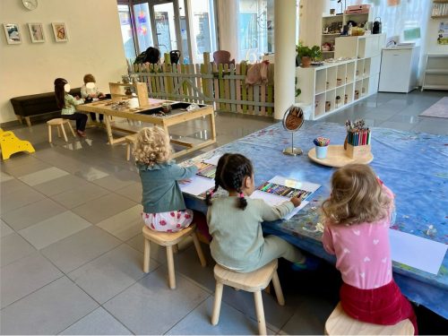 Übersicht der Spielgruppe: Zwei Kinder am sensorischen Tisch und drei Kinder beim Malen am Basteltisch in Zürich.