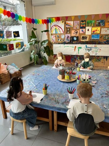 Vier Kinder sitzen am großen Mal- und Basteltisch und arbeiten kreativ, im Hintergrund ist die Malwand der Spielgruppe in Zürich zu sehen.